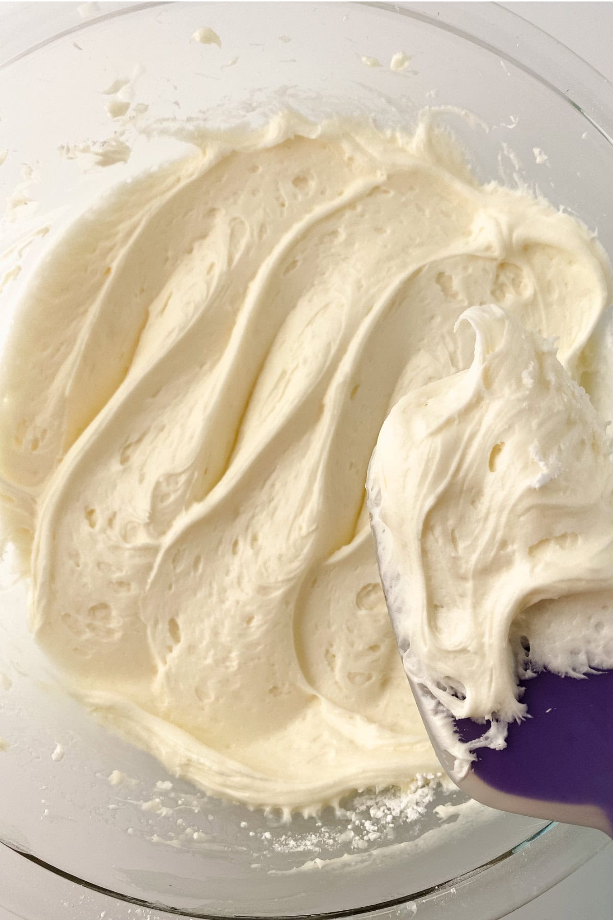 A glass bowl filled with creamy, whipped cream cheese frosting. A purple spatula is scooping up some of the smooth, fluffy mixture.