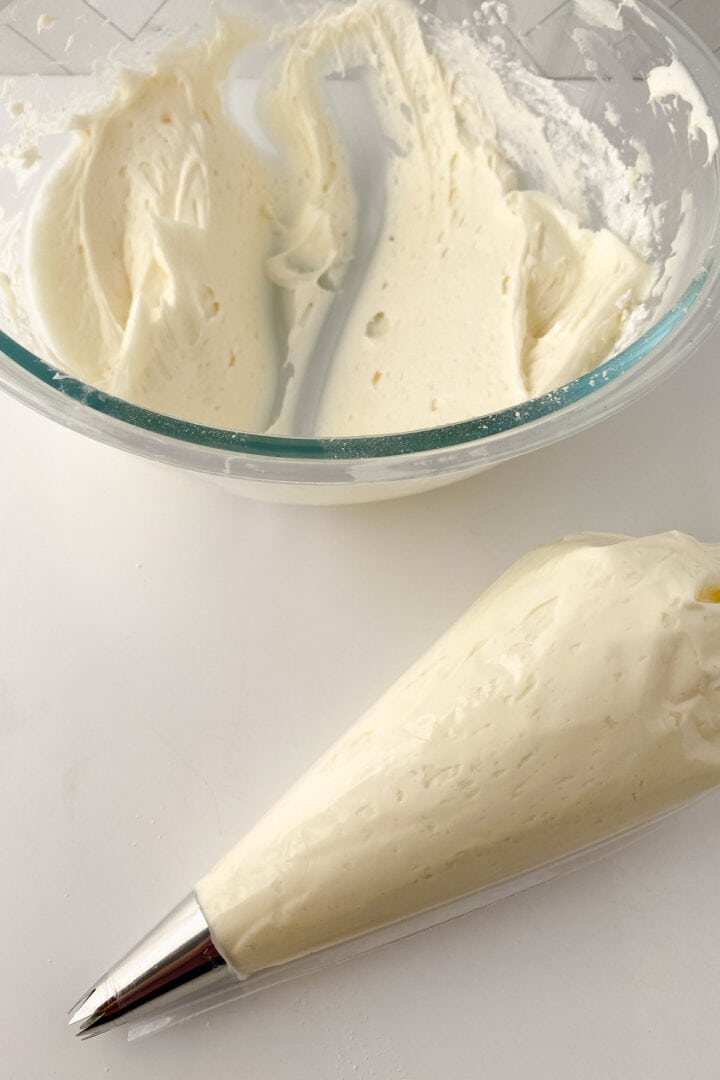 A glass bowl with creamy white cream cheese frosting sits on a white surface. In front, a piping bag filled with the same frosting and fitted with a metal tip lies ready for use.