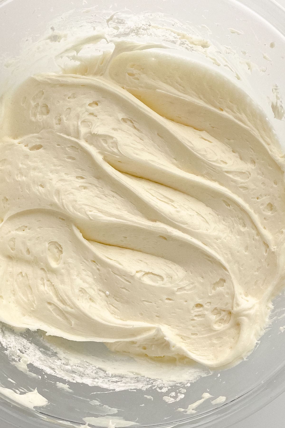A close-up of creamy, smooth cream cheese frosting swirled in a mixing bowl, showing a light, fluffy texture with soft peaks and ridges.