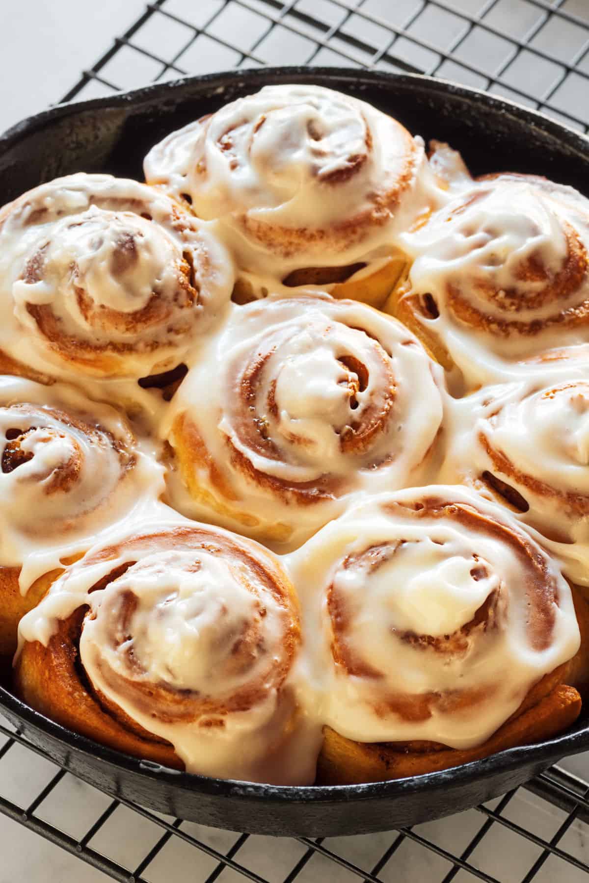 A pan filled with freshly baked cinnamon rolls topped with creamy cream cheese frosting sits on a wire cooling rack. The golden brown, swirled rolls have rich frosting melting over the tops.