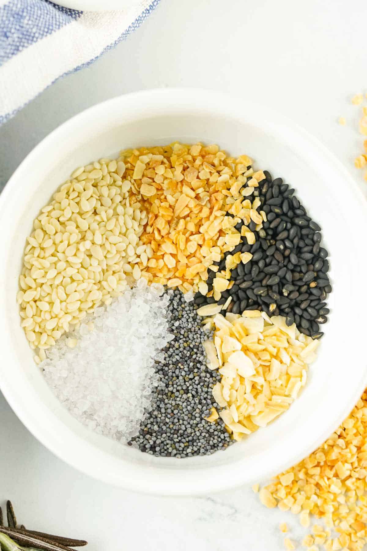A white bowl containing six types of ingredients arranged in sections: white sesame seeds, dried minced onion, black sesame seeds, dried minced garlic, poppy seeds, and coarse salt-classic components of an everything bagel seasoning blend.