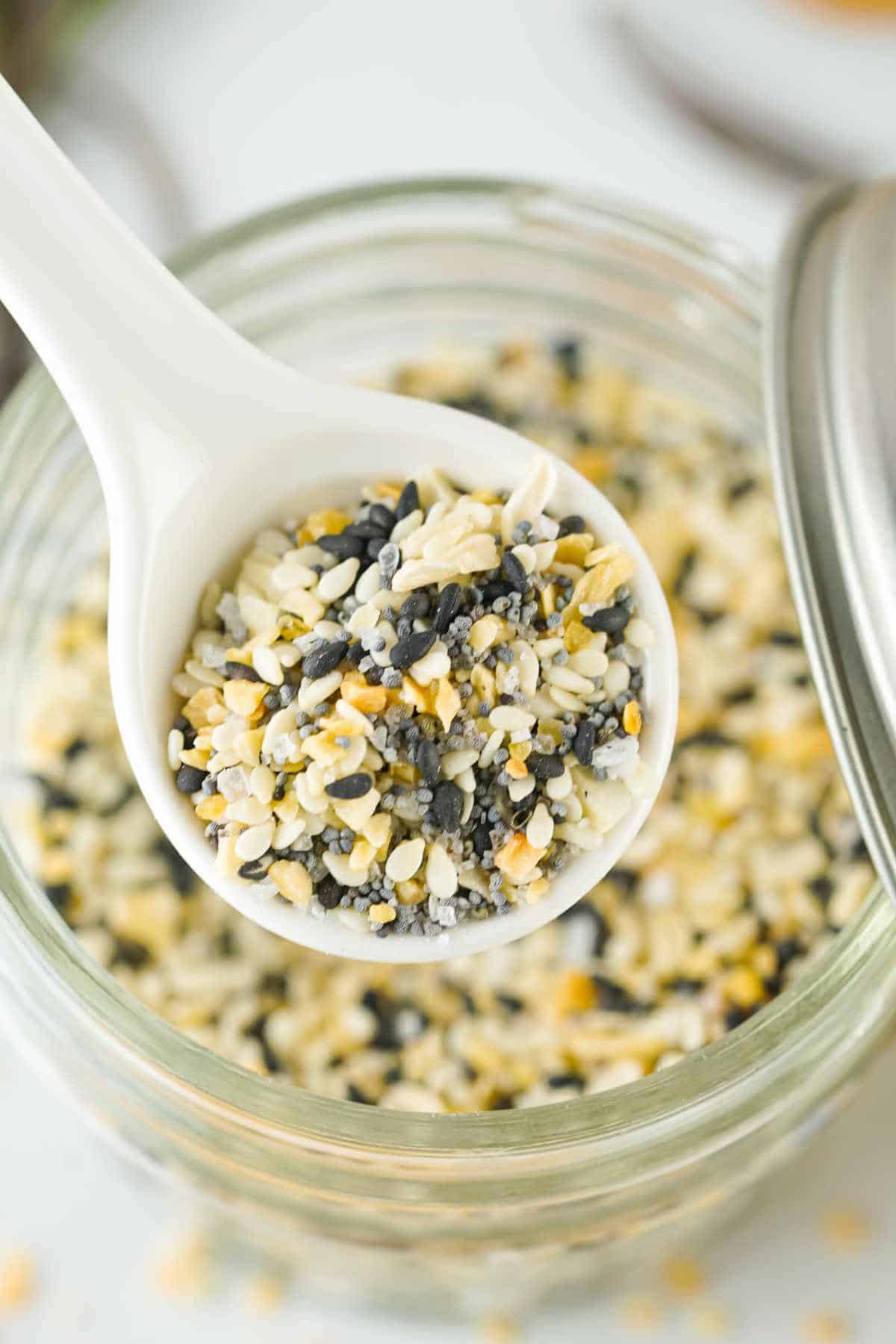 A white spoon holds everything bagel seasoning blend—sesame seeds, poppy seeds, dried garlic, dried onion, and salt—over an open glass jar filled with the same flavorful mixture.