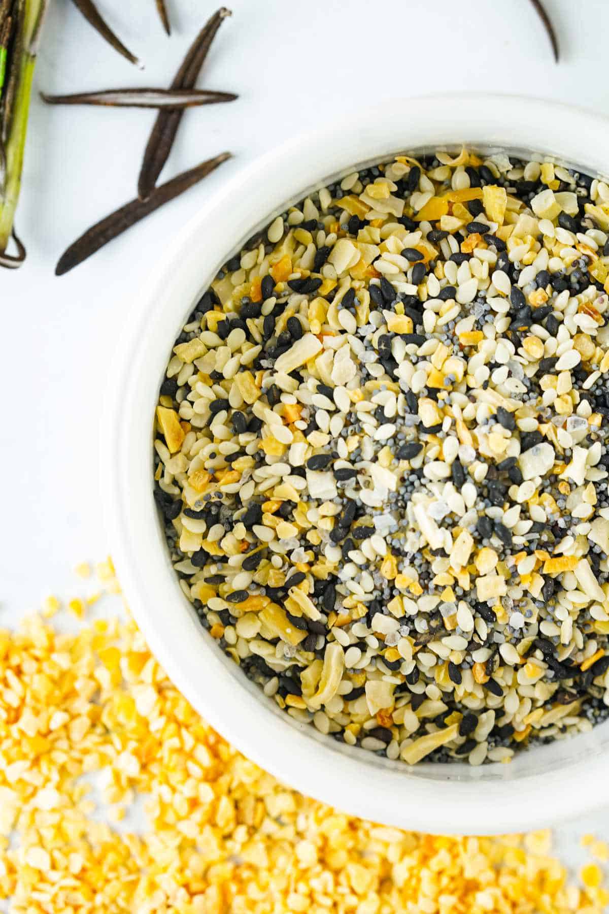 A white bowl filled with everything bagel seasoning blend-sesame seeds, poppy seeds, dried garlic, dried onion, and salt-sits on a white surface with seed pods and scattered bits of the mix nearby.