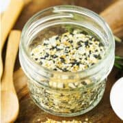 A small glass jar filled with an everything bagel seasoning blend, showing a mix of sesame seeds, poppy seeds, dried garlic, and onion. The jar is on a wooden surface with wooden utensils nearby.