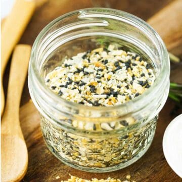 A small glass jar filled with an everything bagel seasoning blend, showing a mix of sesame seeds, poppy seeds, dried garlic, and onion. The jar is on a wooden surface with wooden utensils nearby.