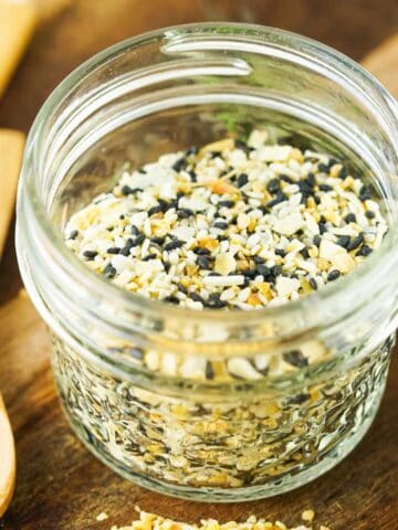 A small glass jar filled with an everything bagel seasoning blend, showing a mix of sesame seeds, poppy seeds, dried garlic, and onion. The jar is on a wooden surface with wooden utensils nearby.