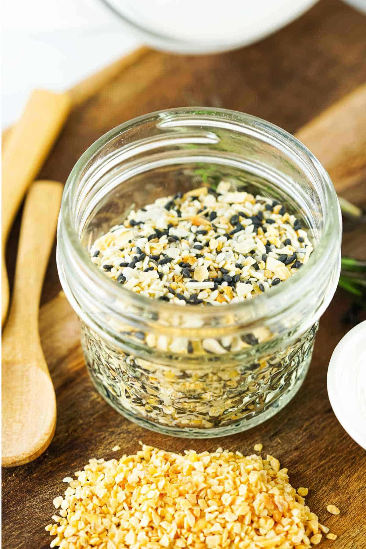 A small glass jar filled with everything bagel seasoning blend sits on a wooden surface, with a wooden spoon nearby and some loose seasoning blend spilled in front of the jar.