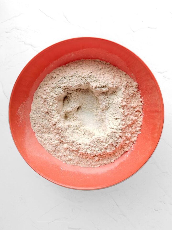 A red bowl filled with flour, ready for making whole wheat flour tortillas, sits with a well in the center on a white textured surface.
