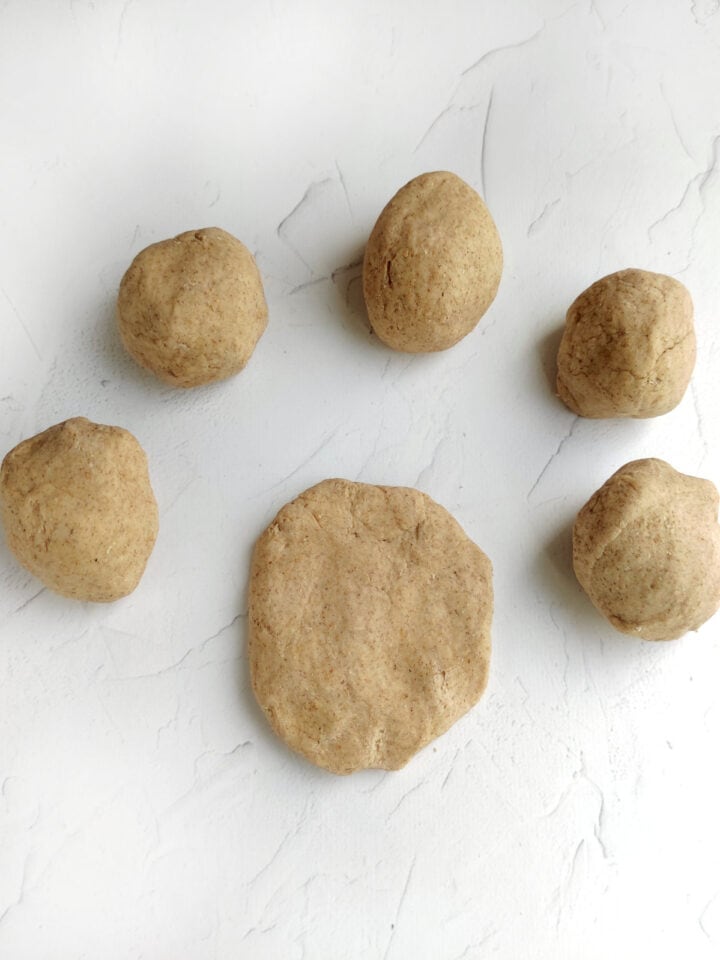 Five pieces of brown dough, perfect for whole wheat flour tortillas, are arranged on a white textured surface; four are ball-shaped, and one is flattened in the center.