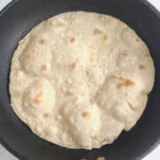 A round, lightly browned whole wheat flour tortilla cooks in a black nonstick pan on a white surface. The tortilla has puffed spots and uneven edges.