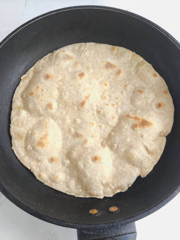 A round, lightly browned whole wheat flour tortilla cooks in a black nonstick pan on a white surface. The tortilla has puffed spots and uneven edges.