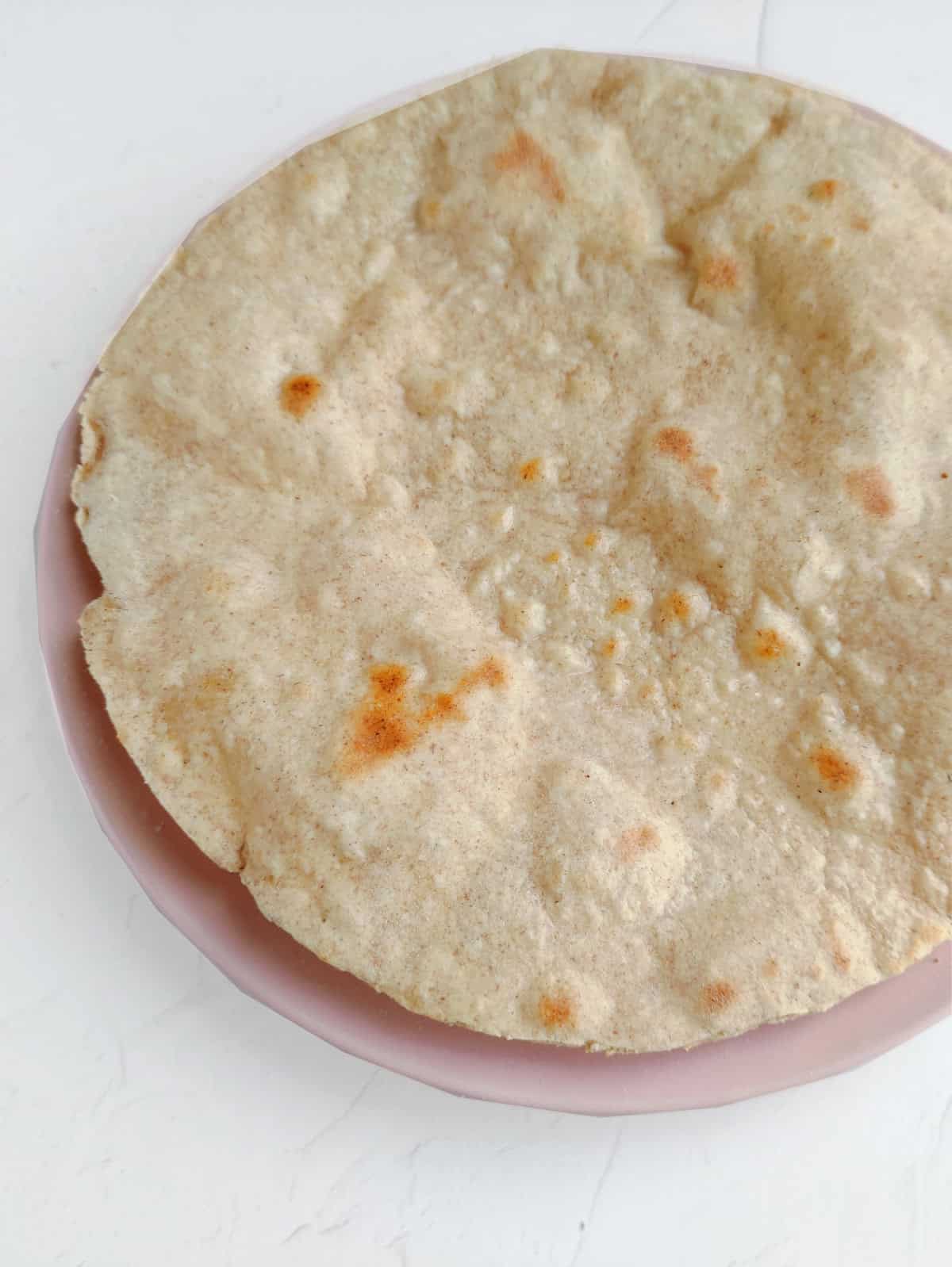 A whole wheat chapati, reminiscent of whole wheat flour tortillas, with light brown spots sits on a pink plate against a white background.