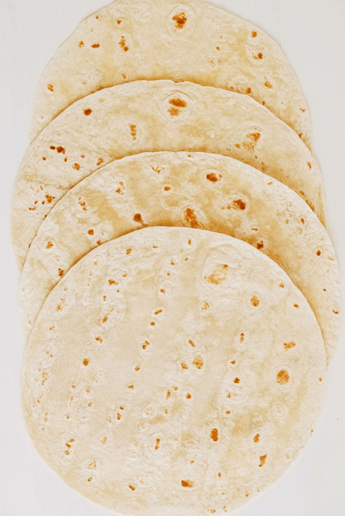 Five whole wheat flour tortillas are stacked in an overlapping pattern on a white surface. The tortillas are round, flat, and lightly browned with small spots.