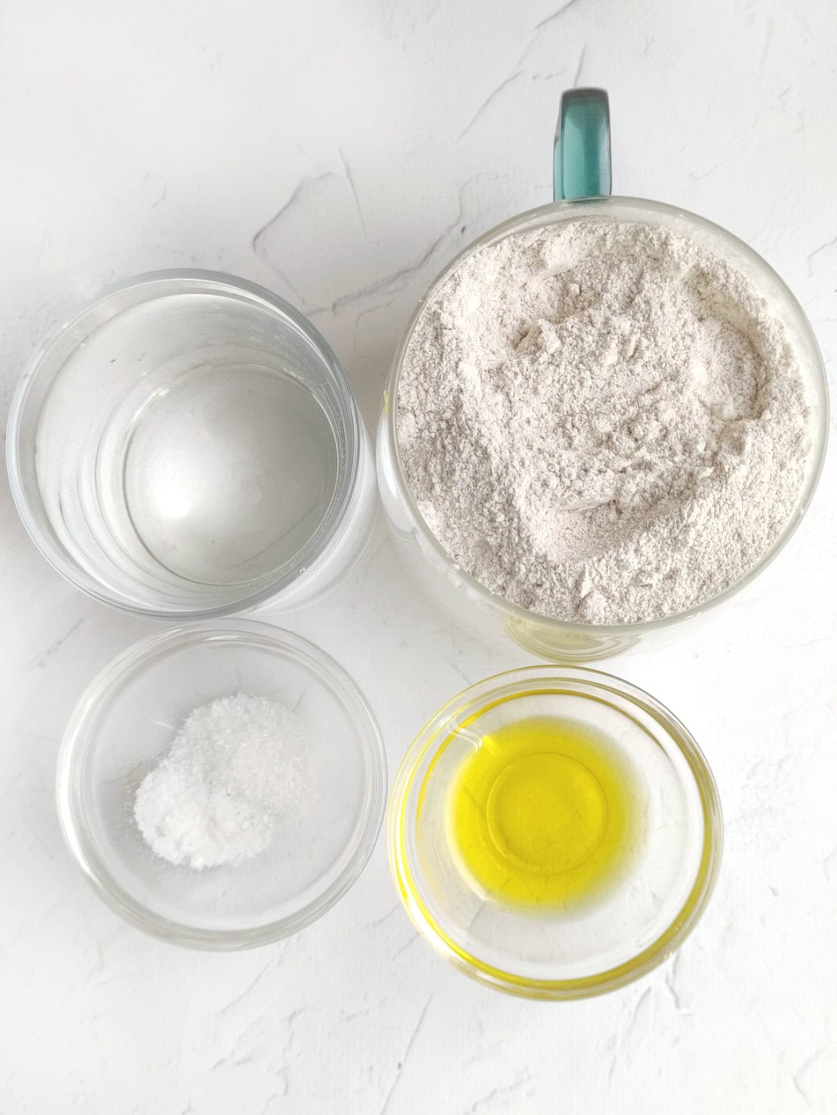 Four glass bowls on a white surface, neatly clustered and filled with water, salt, olive oil, and flour—perfect for making whole wheat flour tortillas from scratch.