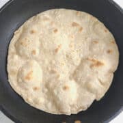 A stack of round, flat whole wheat flour tortillas is arranged on a cork mat atop a white cloth. One tortilla is folded, and a stalk of wheat rests on top, emphasizing the connection to wheat flour.