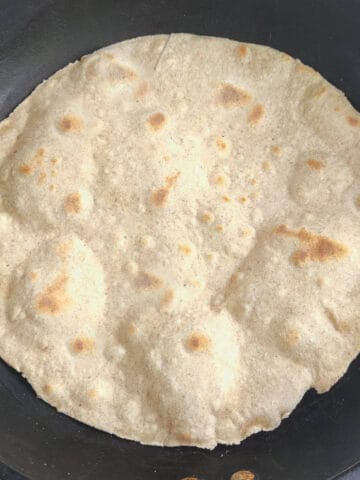 A stack of round, flat whole wheat flour tortillas is arranged on a cork mat atop a white cloth. One tortilla is folded, and a stalk of wheat rests on top, emphasizing the connection to wheat flour.