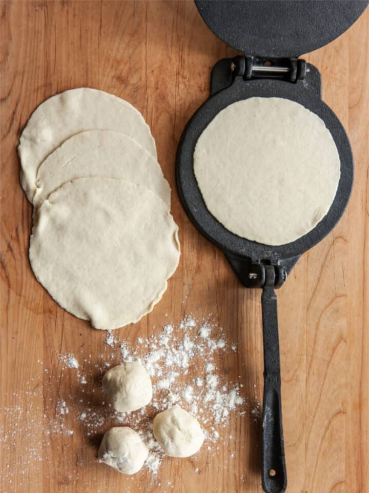 A tortilla press with a round piece of dough sits on a wooden surface next to three rolled dough balls, a dusting of flour, and three flattened, uncooked whole wheat flour tortillas.