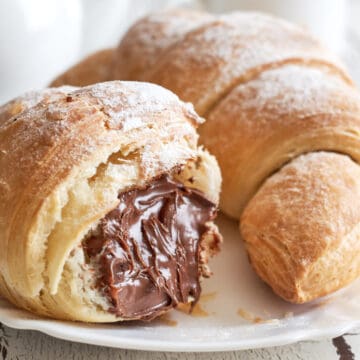 Two flaky croissants on a white plate, one split open to reveal a rich chocolate hazelnut filling, dusted lightly with powdered sugar.