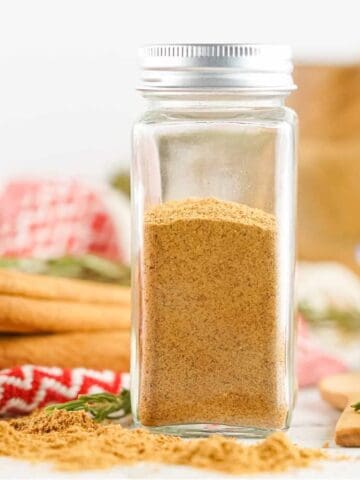 A glass spice jar filled with ground cinnamon sits on a white surface, surrounded by cinnamon sticks, scattered cinnamon powder, and a red and white patterned cloth—perfect for adding to your favorite gingerbread spice mix.
