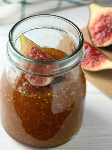 A glass jar filled with honey and fig jam, topped with a fresh fig slice. In the background, fresh figs are cut open and placed on a wooden board. The scene is set on a white surface.