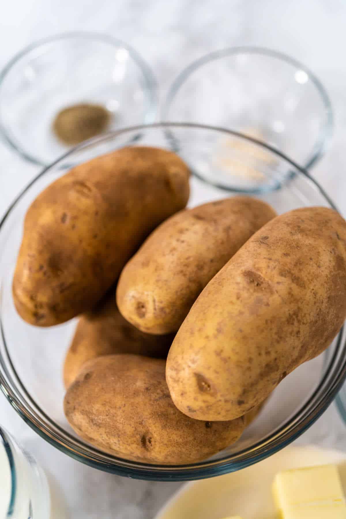 A glass bowl filled with several raw russet potatoes sits on a countertop, ready for making Irish Farls or potato bread, with two small glass bowls containing seasonings and a plate with a few butter slices nearby.