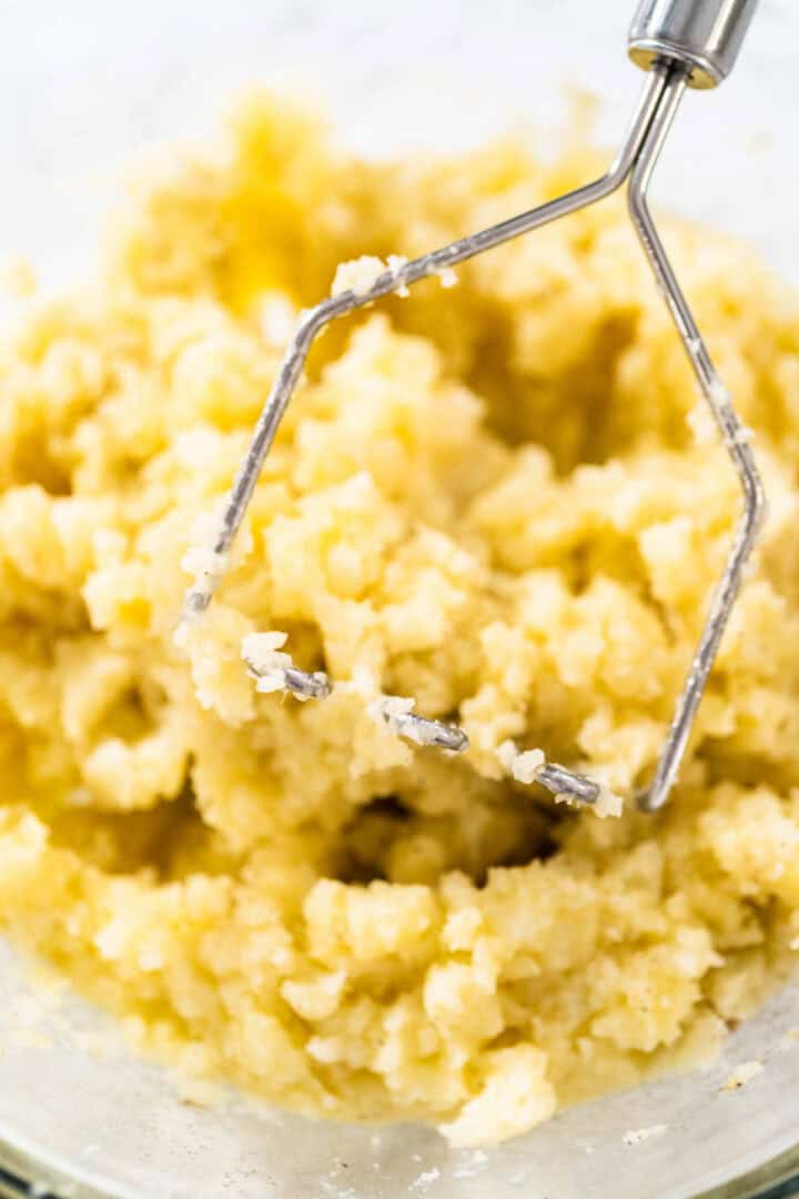A metal potato masher is mashing yellow potatoes in a glass bowl, creating a fluffy, textured mixture—perfect for making Irish potato pancakes or Traditional Irish farls.