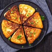 A round plate holds six golden-brown, triangular slices of Irish potato pancakes, garnished with chopped parsley, placed on a dark cloth napkin atop a wooden table.