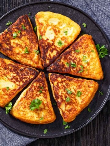 A round plate holds six golden-brown, triangular slices of Irish potato pancakes, garnished with chopped parsley, placed on a dark cloth napkin atop a wooden table.
