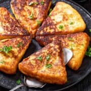 A black plate with six golden-brown, triangular slices of French toast reminiscent of Traditional Irish farls, garnished with fresh parsley, with a serving spoon underneath one slice. The background shows a rustic wooden table.