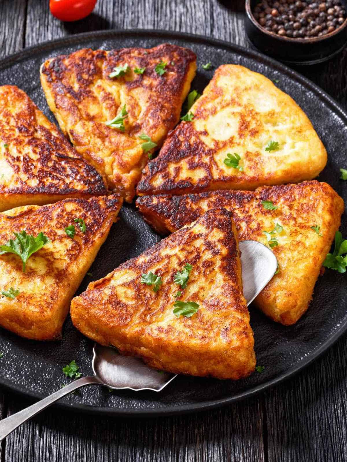 A black plate with six golden-brown, triangular slices of French toast reminiscent of Traditional Irish farls, garnished with fresh parsley, with a serving spoon underneath one slice. The background shows a rustic wooden table.