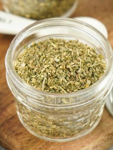 A small glass jar filled with a classic Italian Seasoning blend sits on a wooden surface, accompanied by a metal lid and a white measuring spoon.