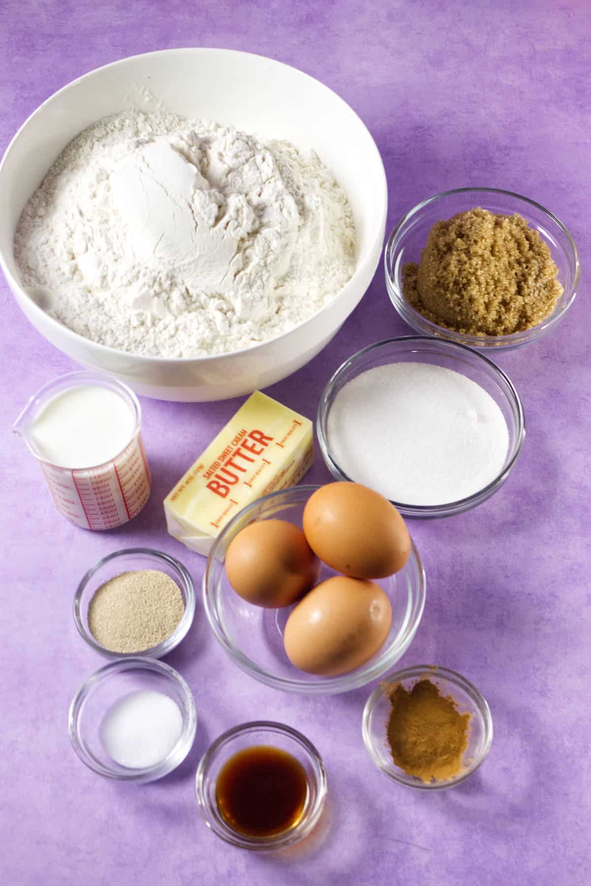 A variety of baking ingredients for Mardi Gras king cake on a purple surface, including a large bowl of flour, brown sugar, granulated sugar, milk, butter, eggs, yeast, salt, vanilla extract, and ground cinnamon in small bowls.