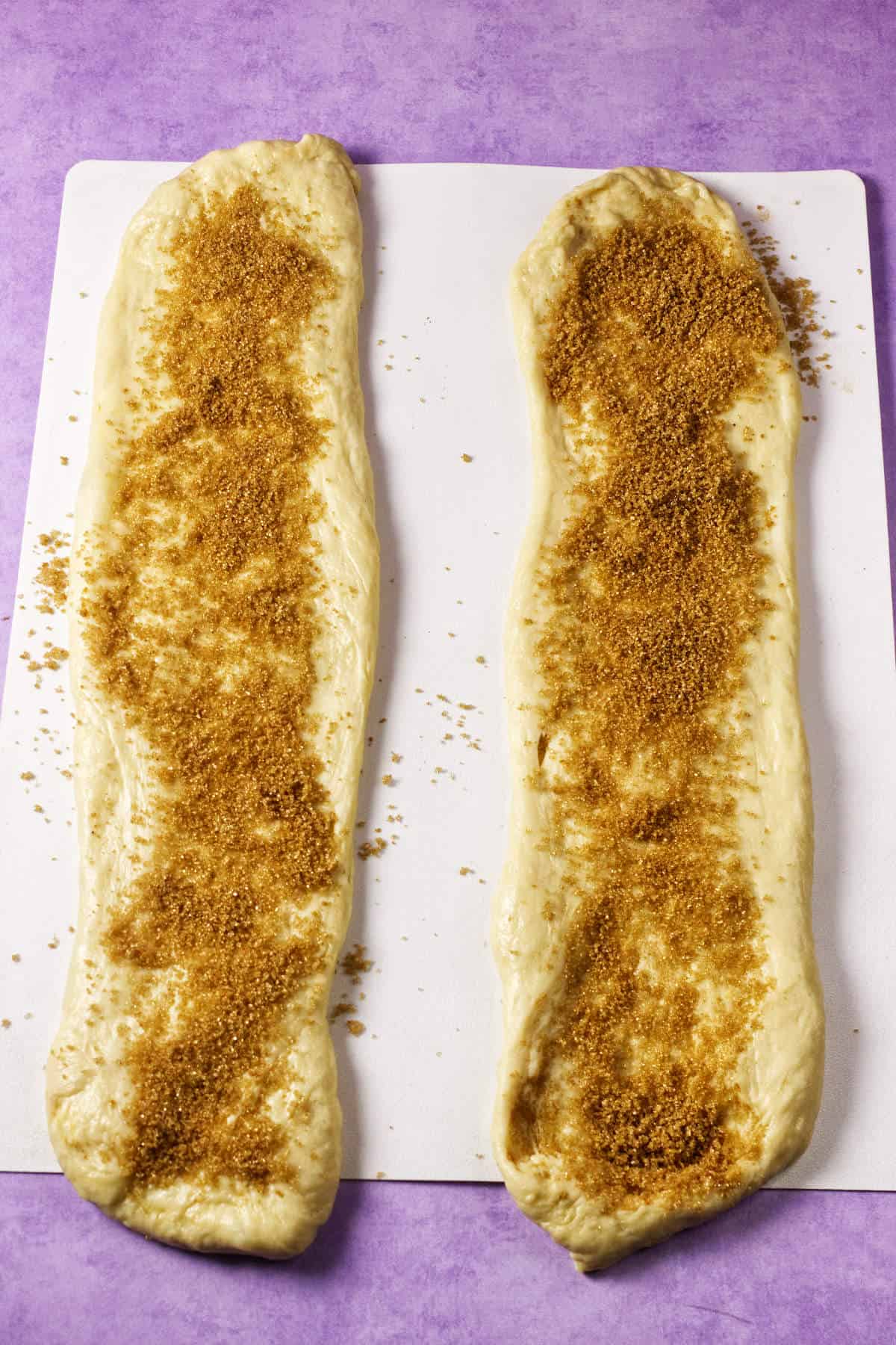 Two elongated strips of dough, reminiscent of mardi gras king cake, are topped with brown sugar and rest on a white cutting board placed on a purple surface.