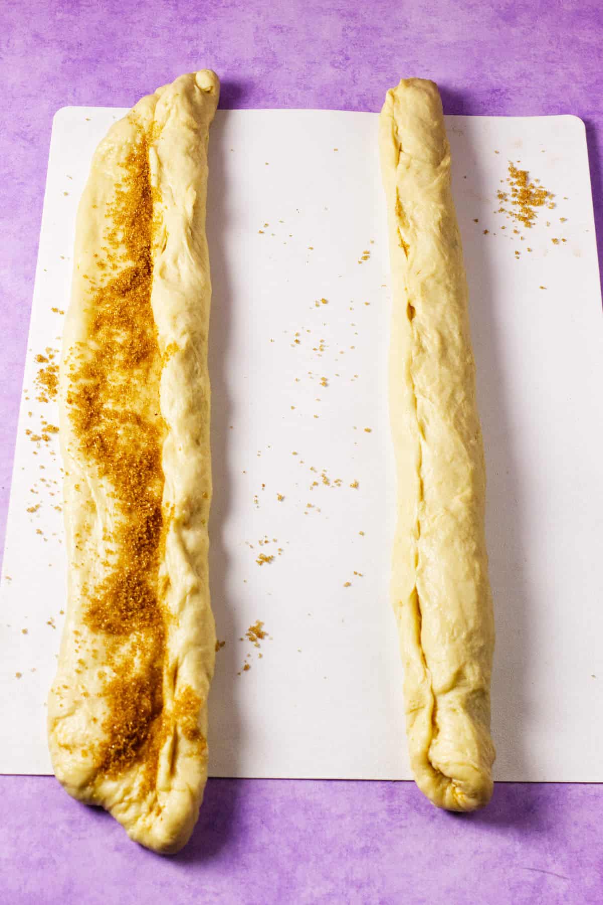 Two strips of dough for a Mardi Gras king cake rest on a white surface over a purple background. The left strip is partially filled with brown sugar and half rolled, while the right strip is fully rolled up.