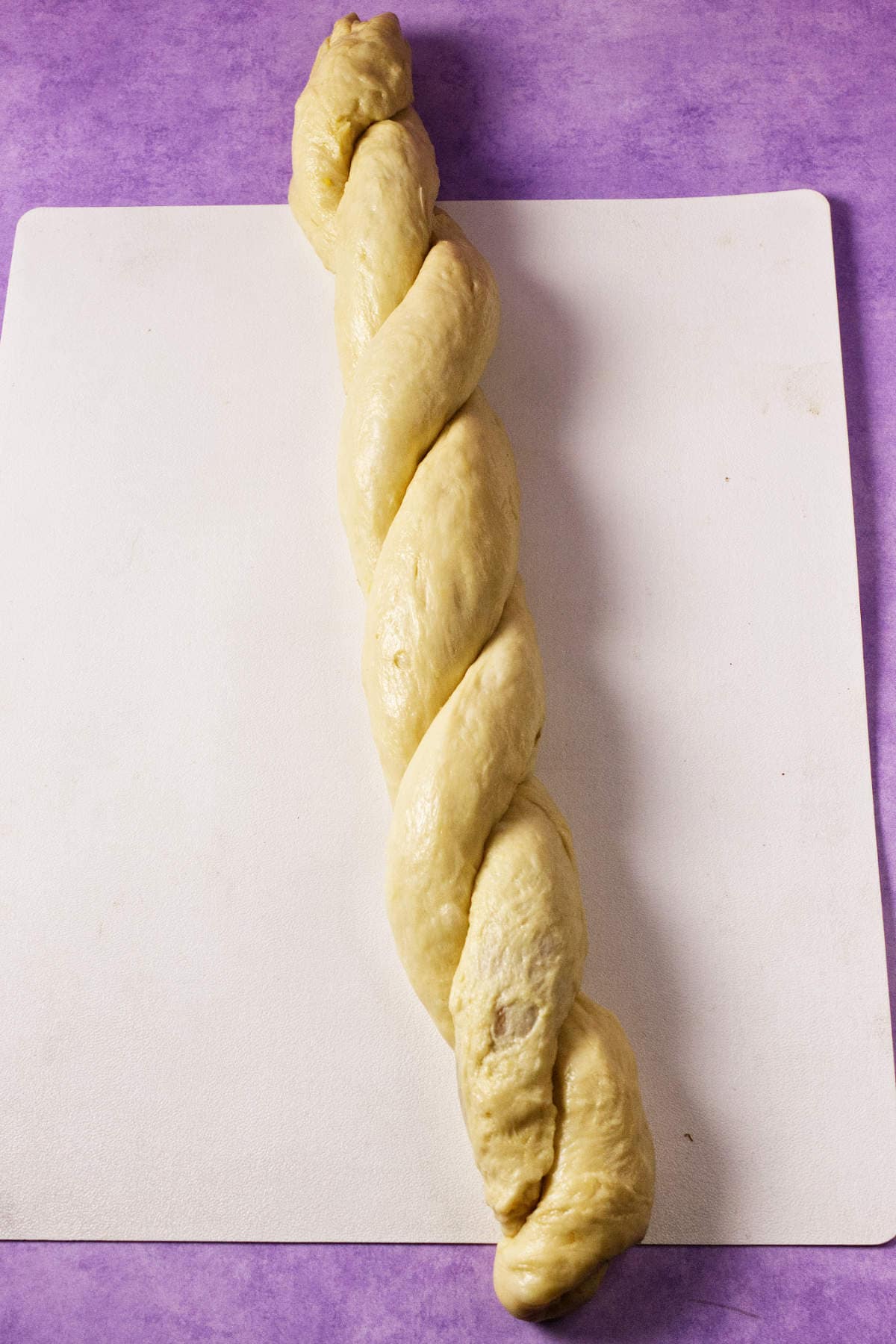A braided loaf of unbaked dough, resembling a Mardi Gras king cake, rests on a white cutting board set against a purple surface.