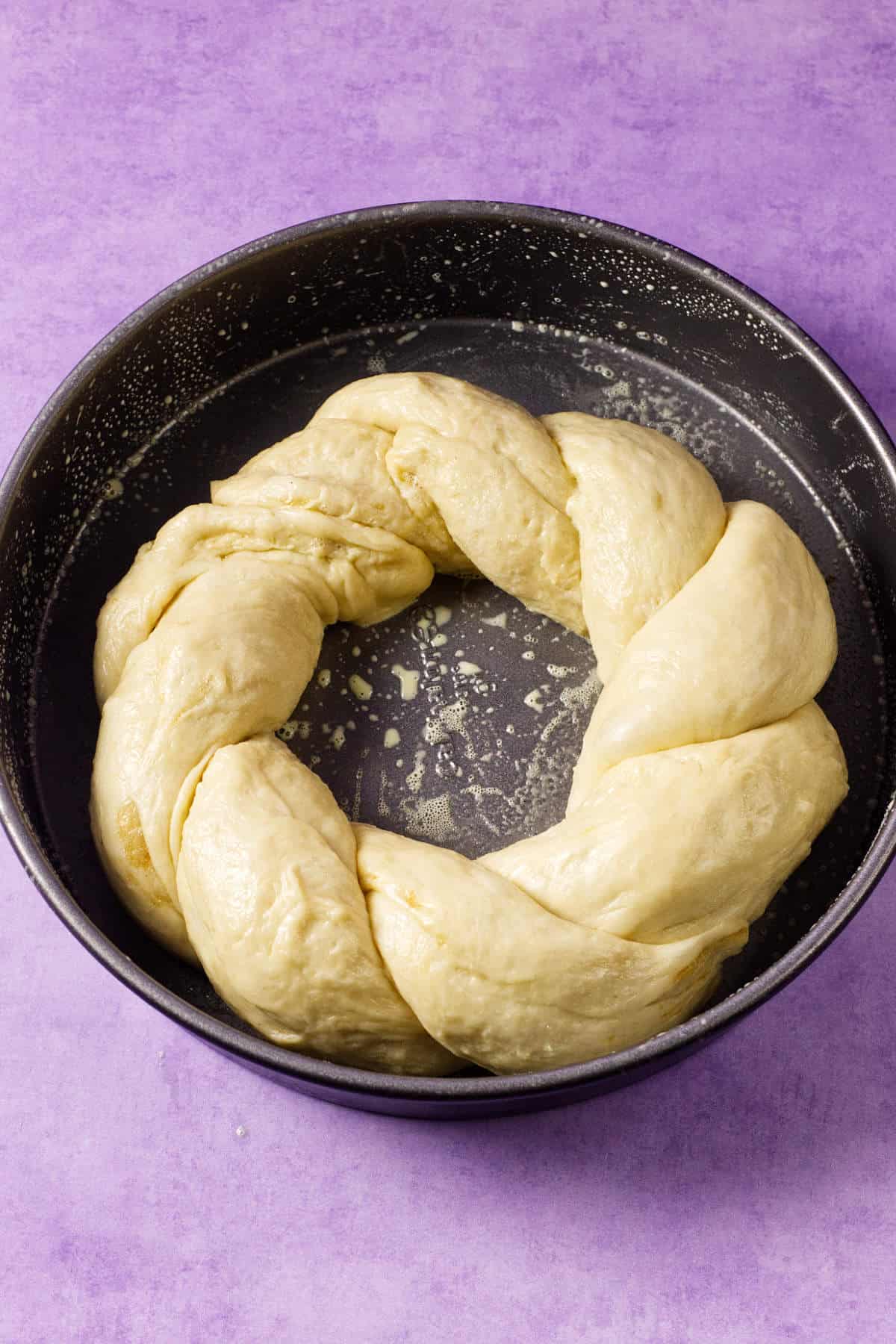 A round baking pan holds a ring of braided, unbaked mardi gras king cake dough on a light purple surface. The thick, slightly glossy dough is ready for baking.
