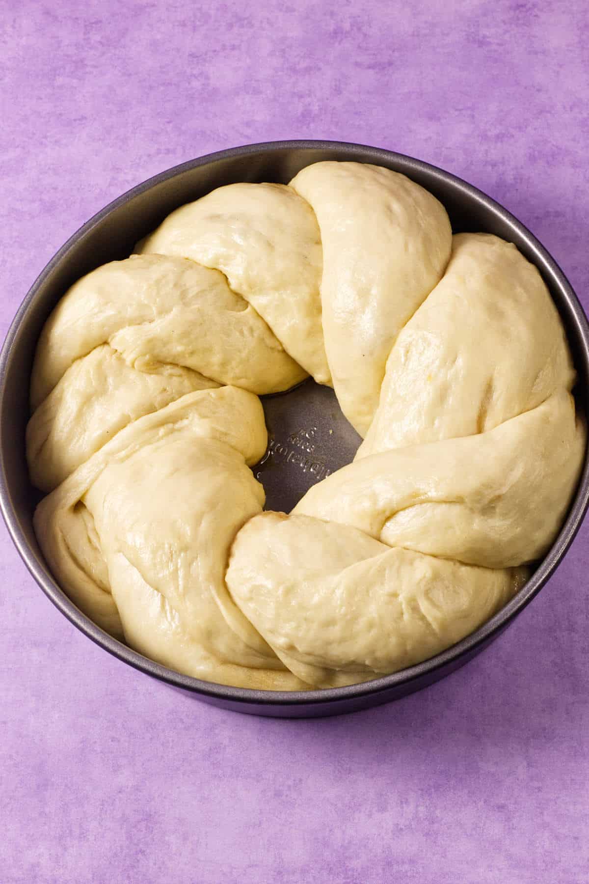 A round baking pan holds braided, unbaked dough arranged in a circle, ready to become a festive mardi gras king cake on a light purple surface.
