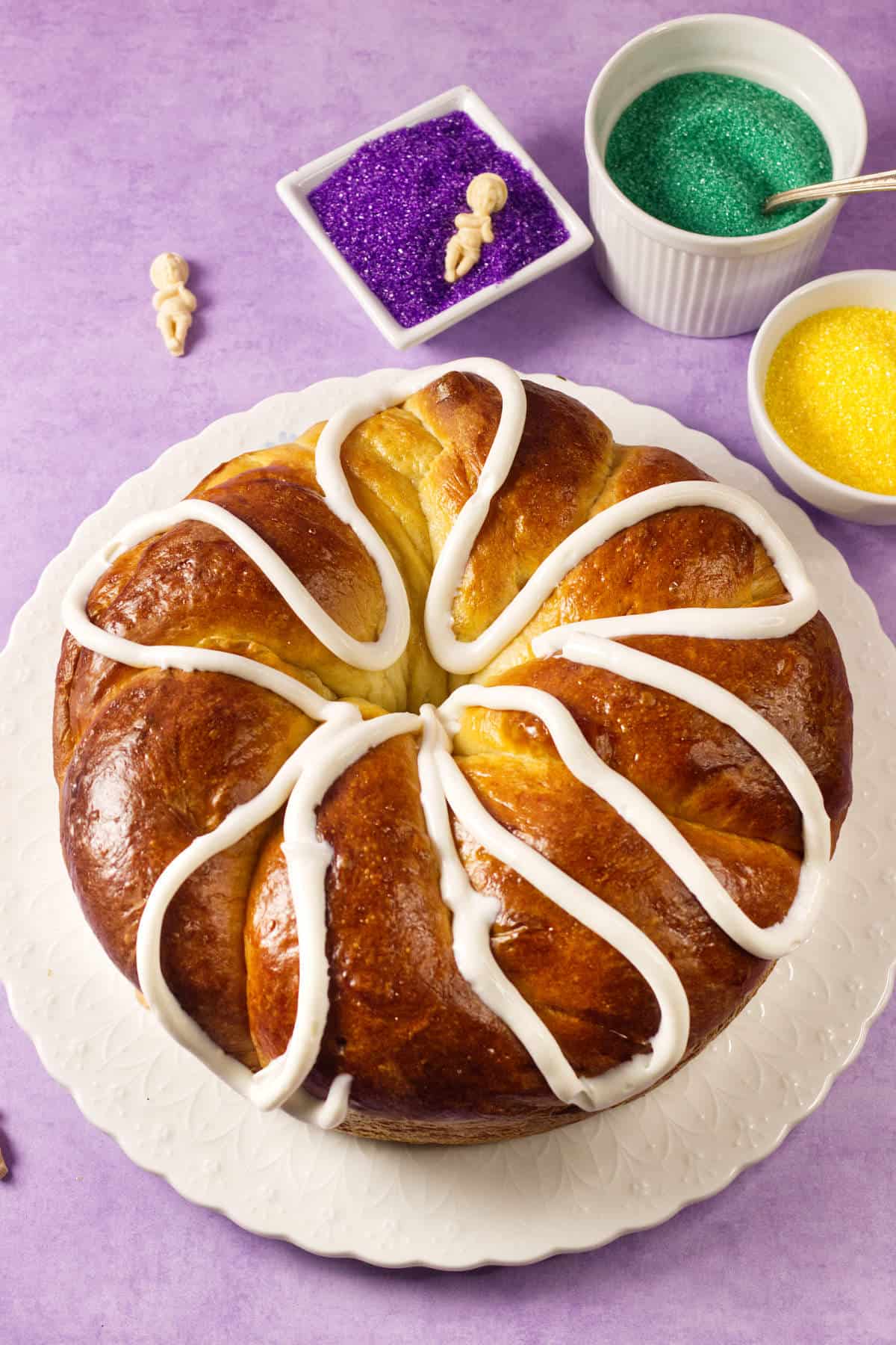 A round Mardi Gras King Cake with white icing drizzled on top sits on a white plate. In the background are small bowls of purple, green, and yellow sugar sprinkles on a light purple surface.