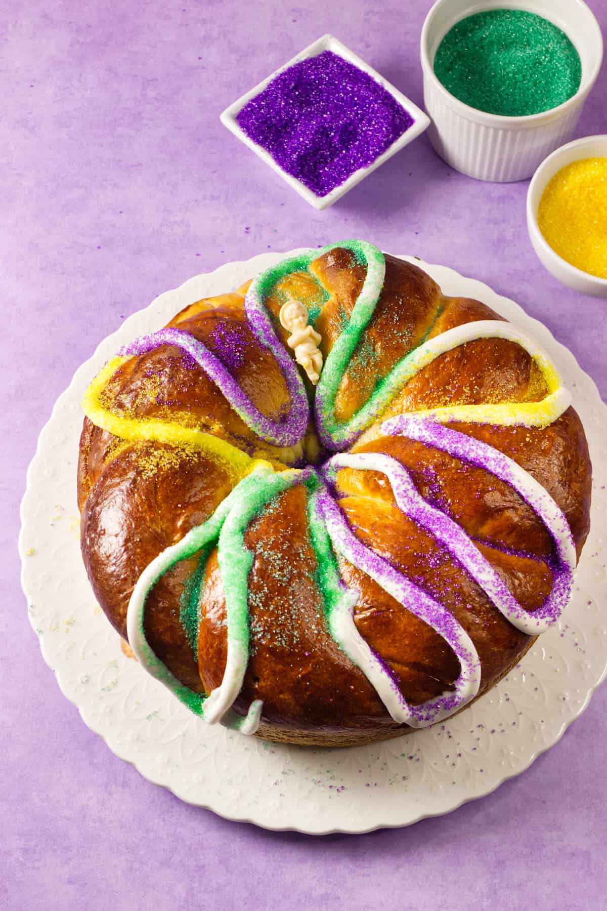 A round Mardi Gras king cake decorated with white icing and sprinkled with yellow, green, and purple sugar sits on a white plate. Bowls of colored sugar are nearby, all against a festive purple background.