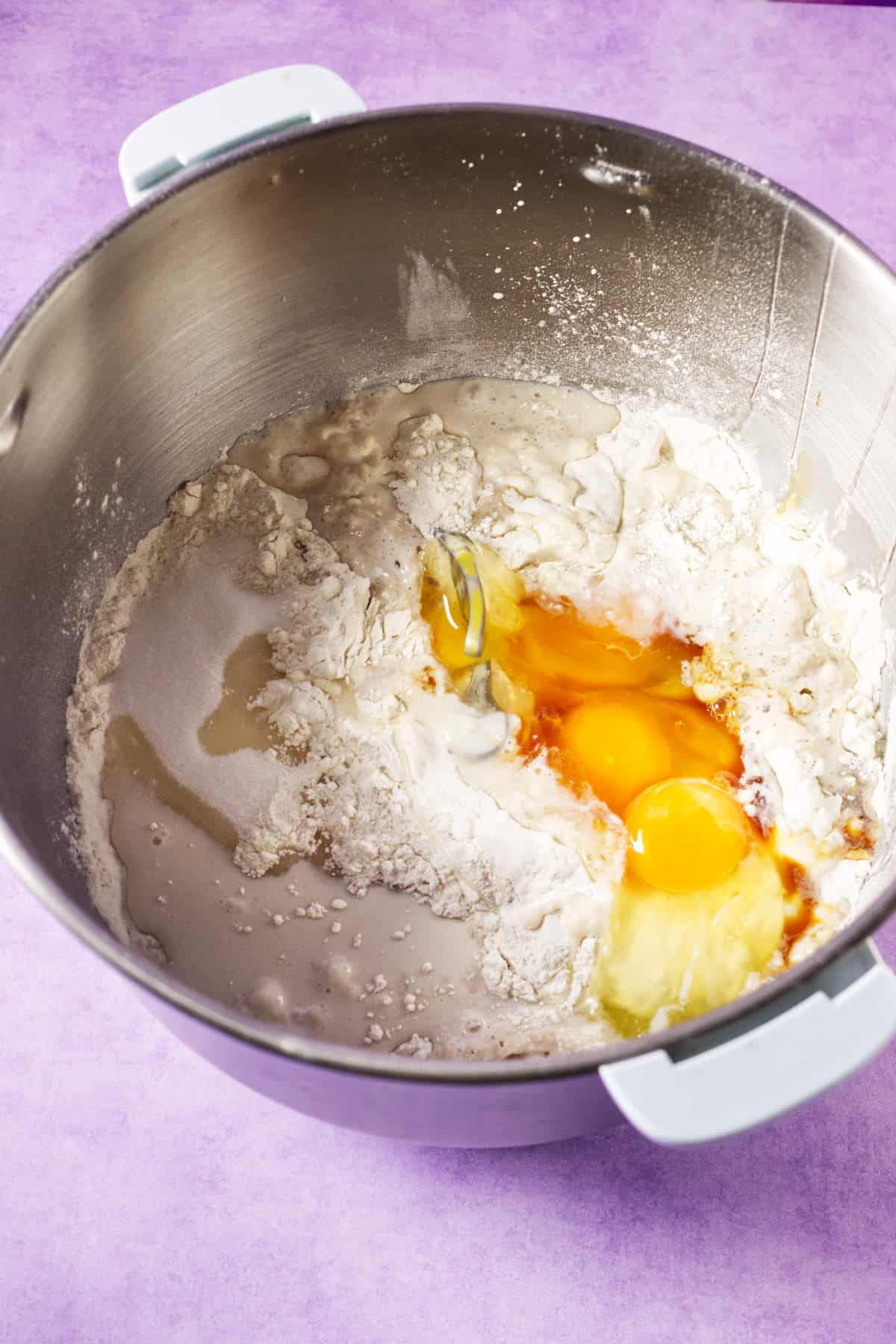 A metal mixing bowl containing flour, two cracked eggs, vanilla extract, and some wet ingredients-this is the start of a festive Mardi Gras king cake, all set to be mixed on a light purple surface.