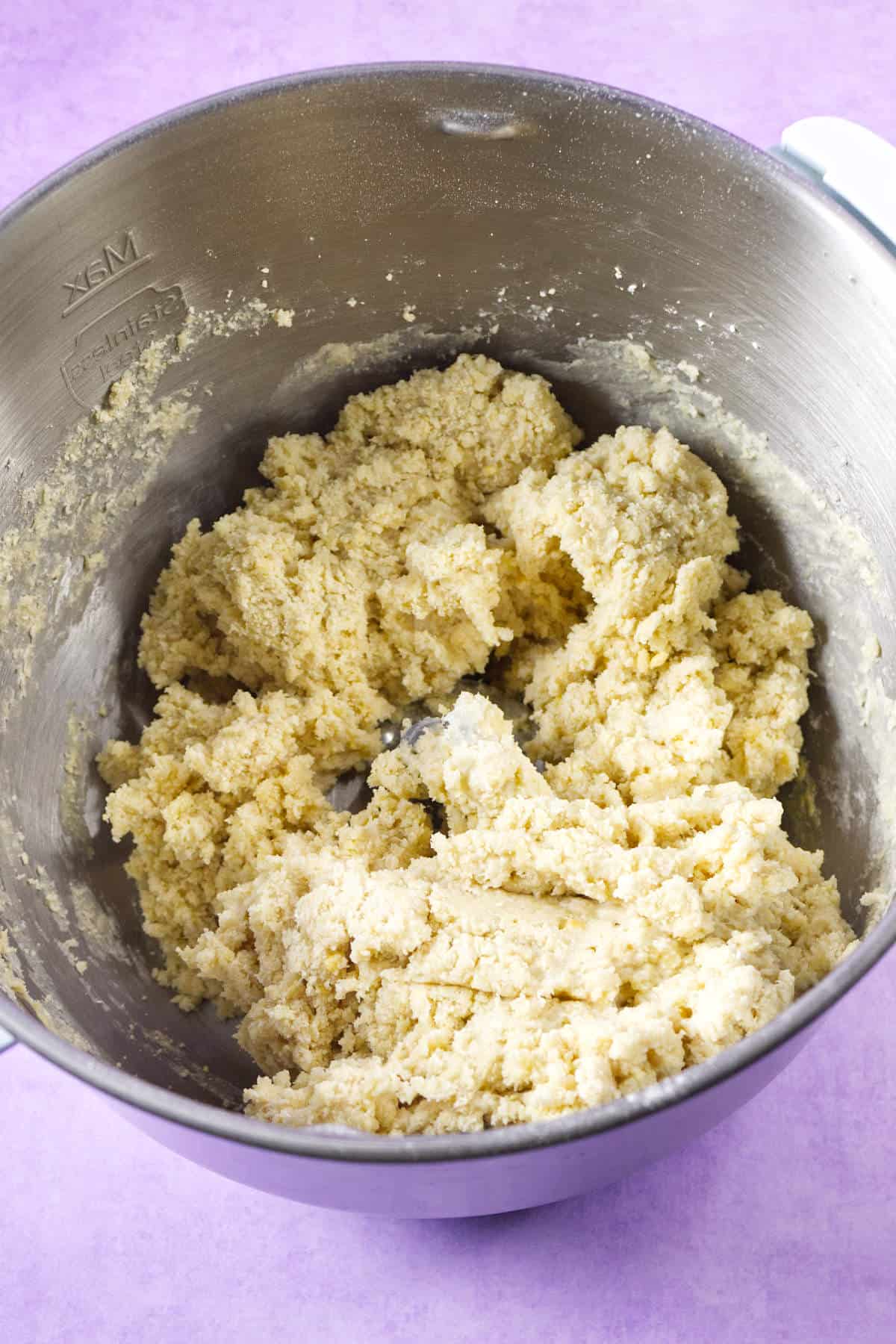 A stainless steel mixing bowl containing raw, crumbly mardi gras king cake cookie dough sits on a light purple surface.