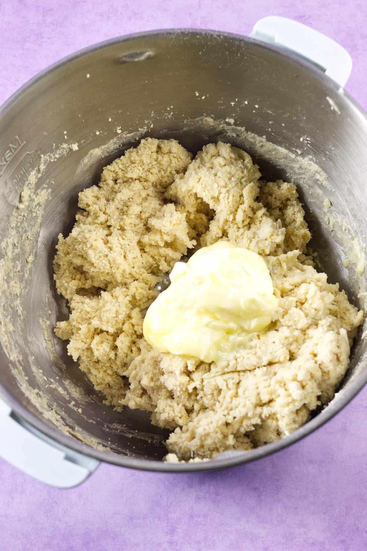 A metal mixing bowl filled with thick cookie dough, topped with a dollop of softened butter, sits on a purple surface-evoking the festive colors of mardi gras king cake.