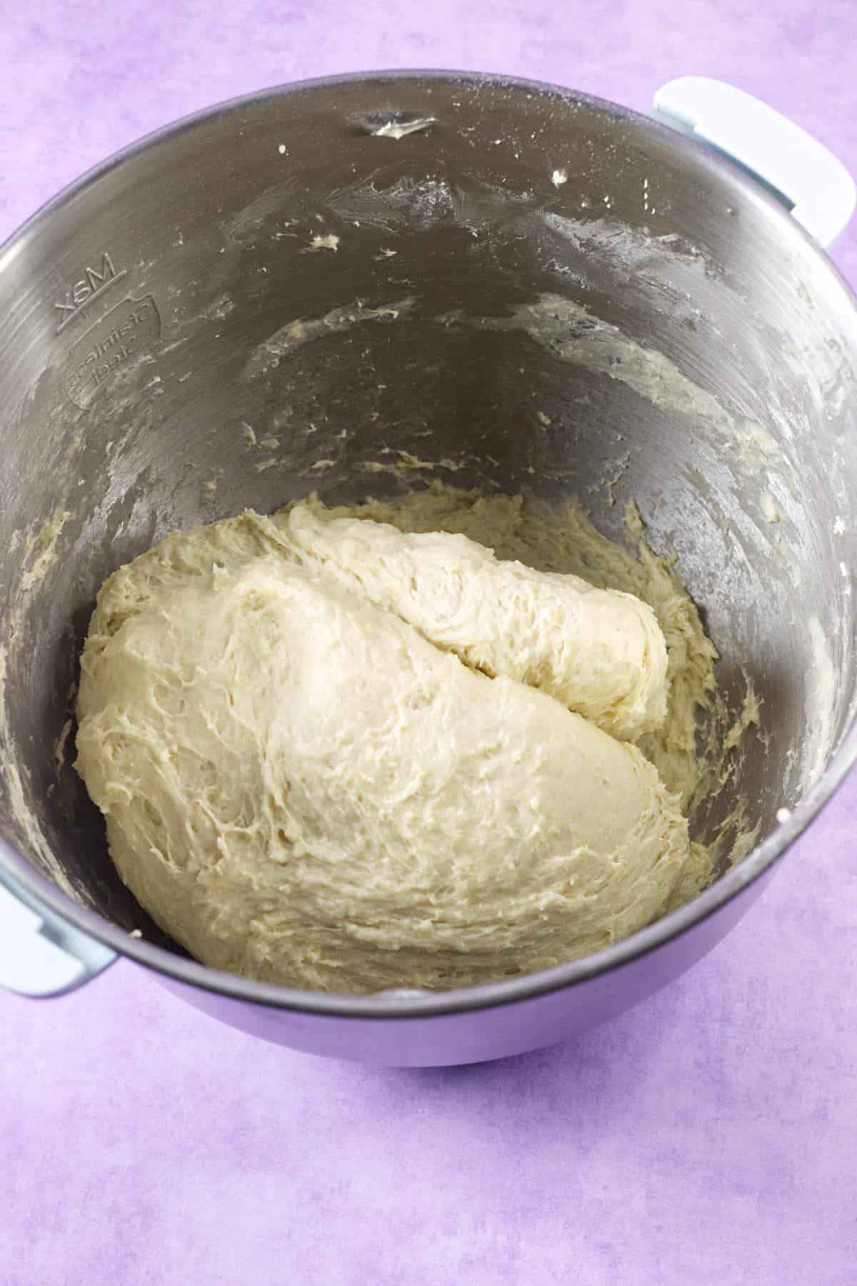 A stainless steel mixing bowl filled with risen, sticky mardi gras king cake dough sits on a purple surface.