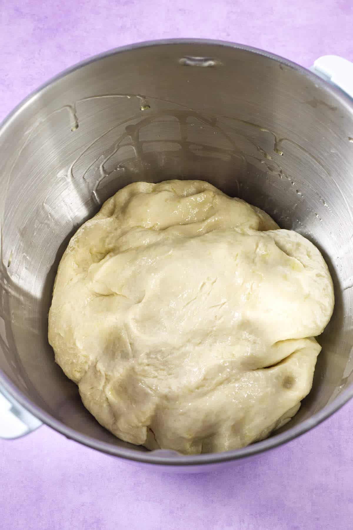 A ball of dough, perfect for a Mardi Gras king cake, rests in a large stainless steel mixing bowl with a light coating of oil. The background features a soft purple surface.