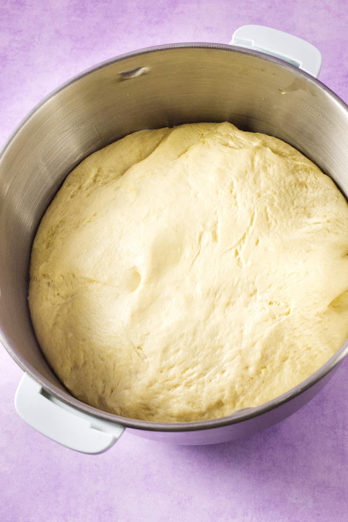 A large stainless steel mixing bowl filled with risen dough for a Mardi Gras king cake, set against a light purple surface.