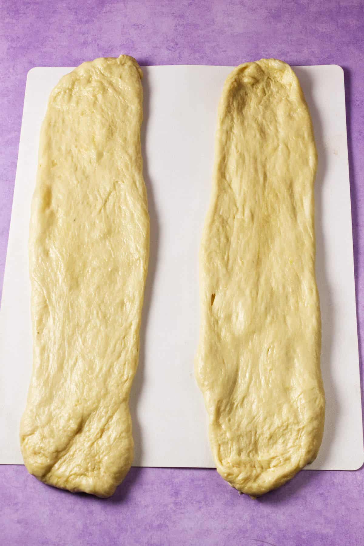 Two rectangular strips of raw dough, ready for a Mardi Gras king cake, are laid side by side on a white surface against a purple background.