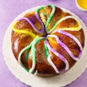 A round Mardi Gras king cake decorated with white icing and sprinkled with yellow, green, and purple sugar sits on a white plate against a purple background.