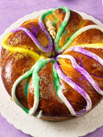 A round Mardi Gras king cake decorated with white icing and sprinkled with yellow, green, and purple sugar sits on a white plate against a purple background.