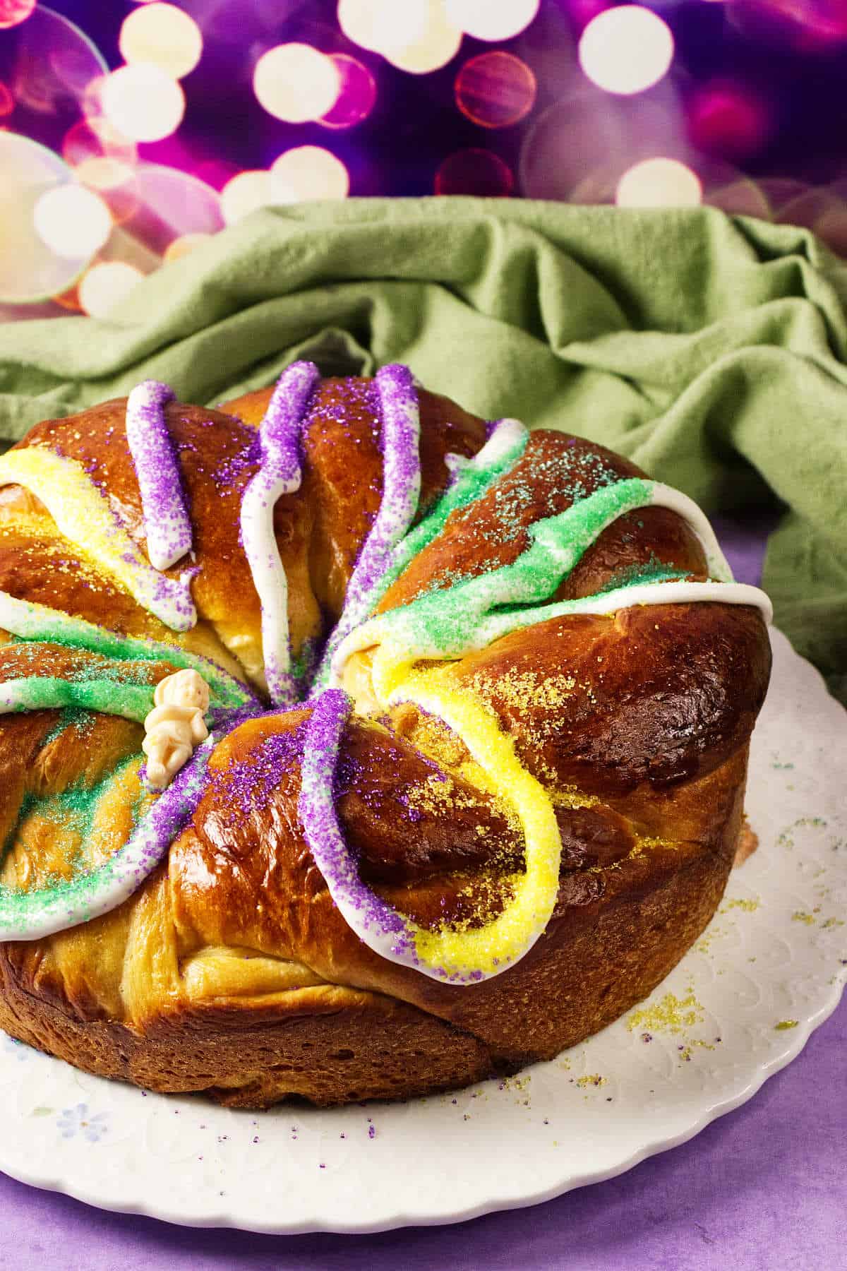 A round Mardi Gras King Cake with colorful sugar toppings in purple, green, and gold, drizzled with white icing, sits on a white plate. A green cloth and festive blurred lights are in the background.