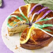 A round Mardi Gras King Cake with colorful sugar and icing in purple, yellow, and green sits on a white plate. One slice is cut out, revealing a small plastic baby inside. A green cloth rests in the background on a purple surface.